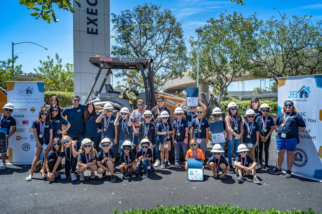 Group of children and adults in hard hats and sunglasses at a Junior Builders event, posing for a group photo outdoors.