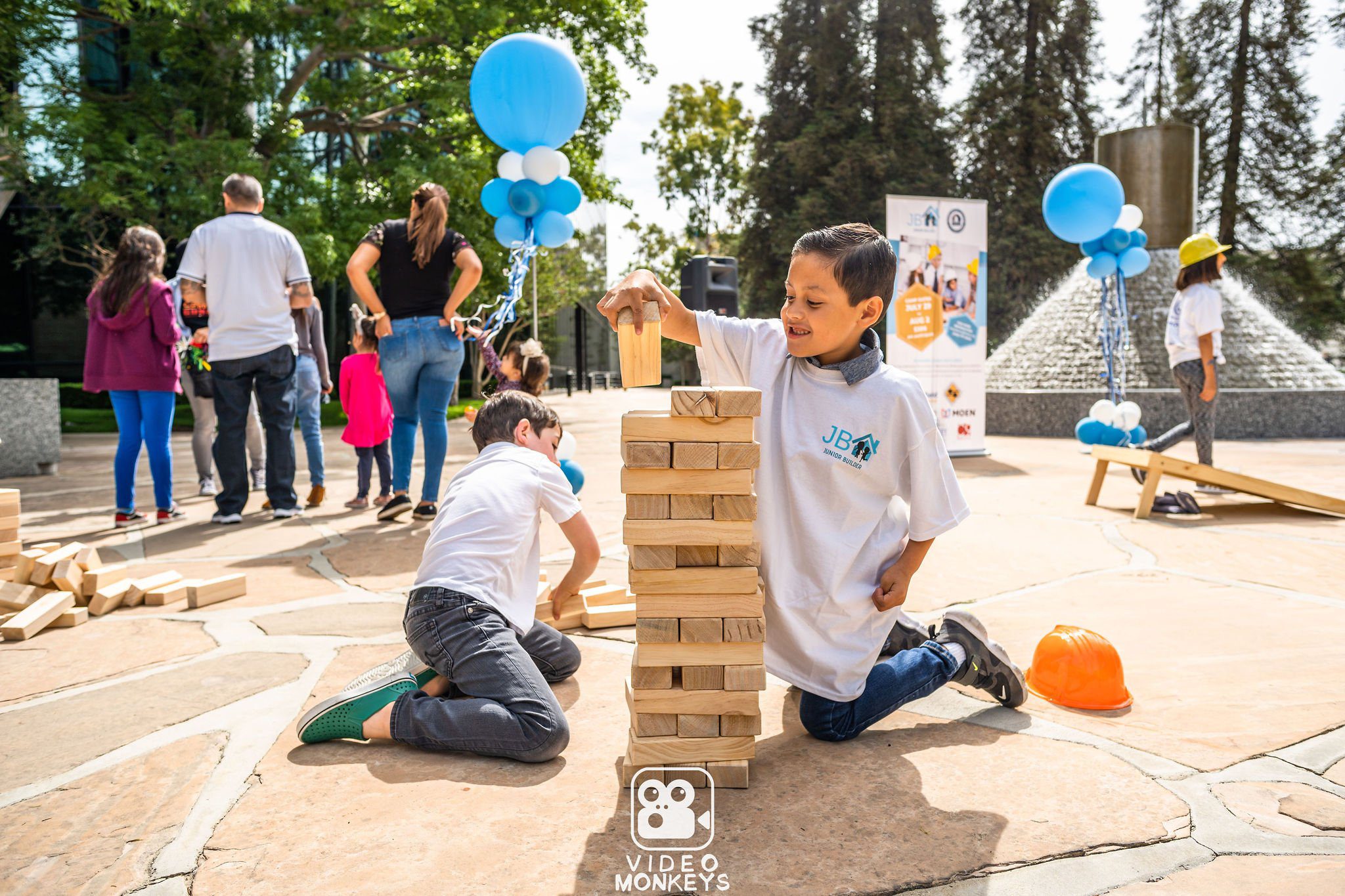 Children stacking large wooden blocks at an outdoor community event, with other people and balloons in the background.