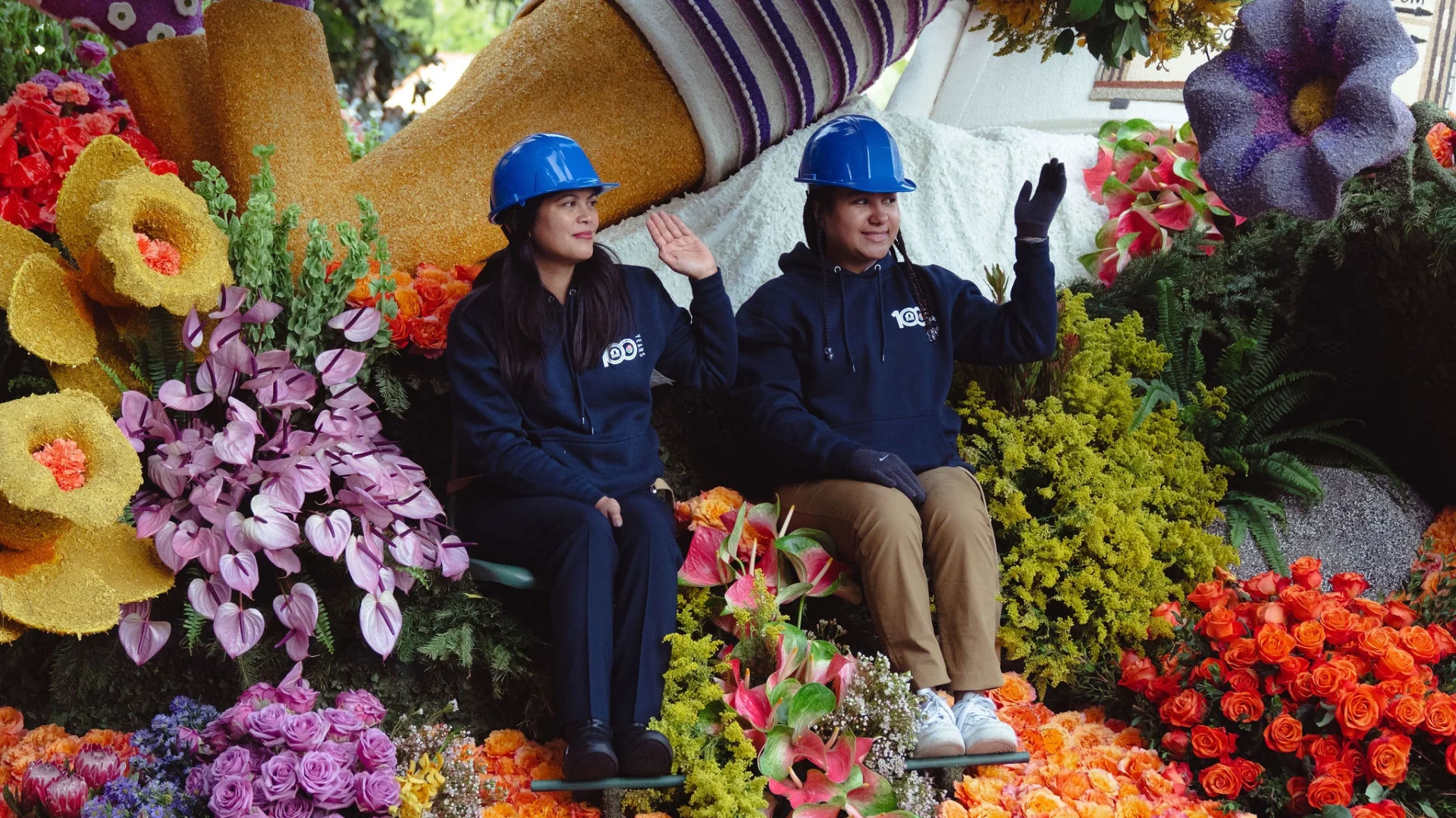 Two Junior Builders wearing blue hard hats and matching hoodies wave while seated on a colorful parade float.