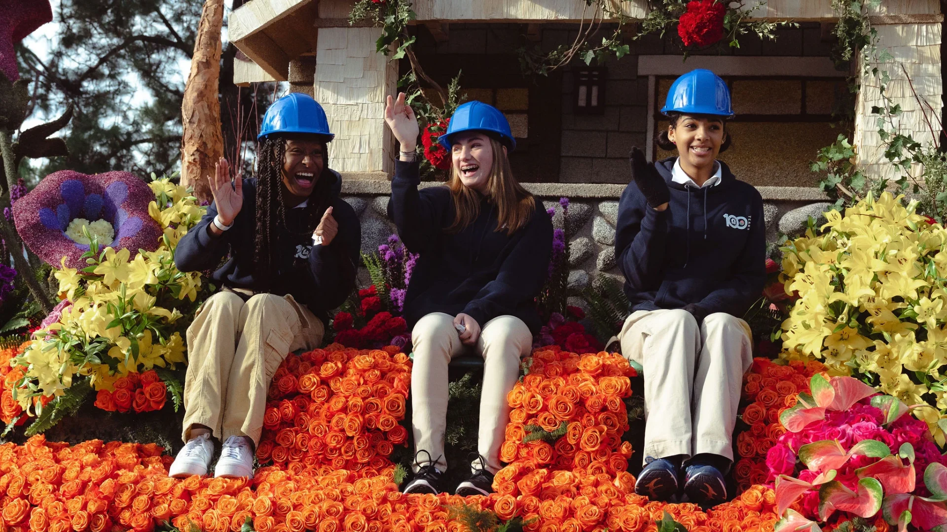 Three women wearing blue hard hats sitting on a flower-covered float, smiling and waving.