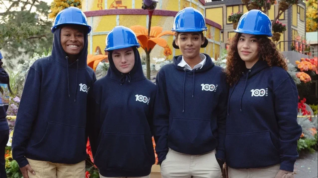 Four people wearing blue hard hats and matching hoodies standing together and smiling in front of a colorful float.